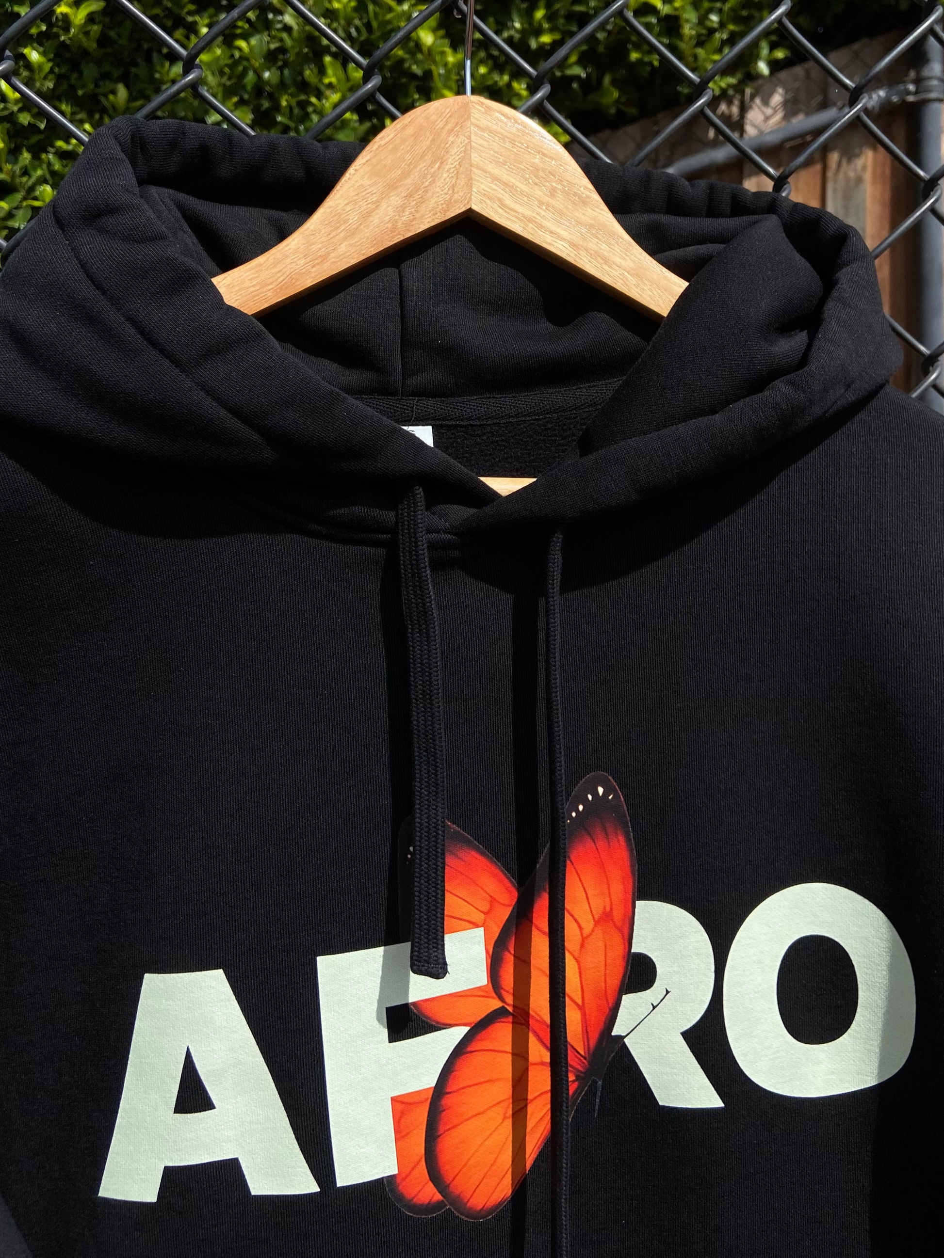 Black hoodie with 'AFRO' and butterfly design on a hanger against a chain-link fence background.